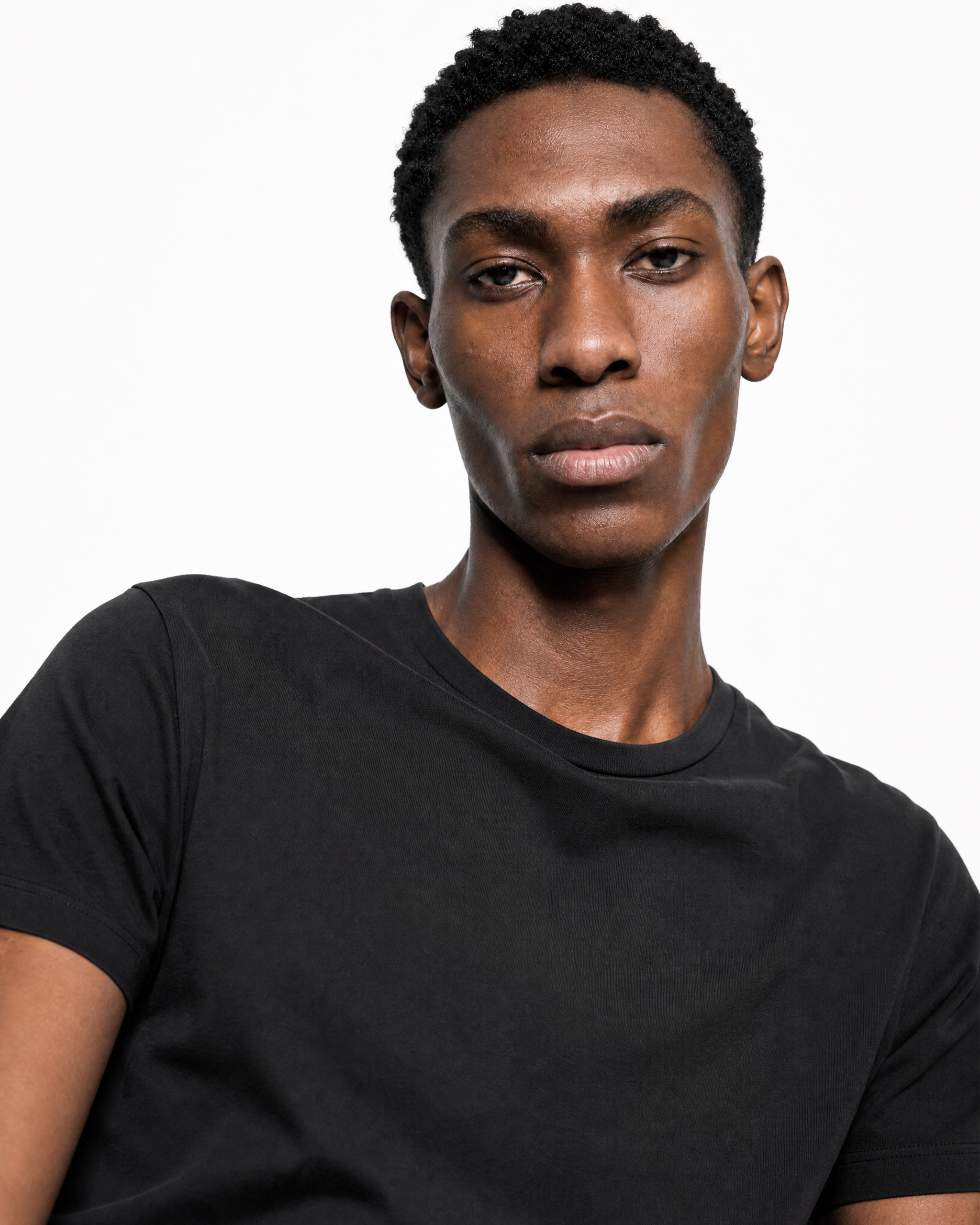 Close-up portrait of a young Black man wearing a black T-shirt, facing the camera with a neutral expression against a white background.