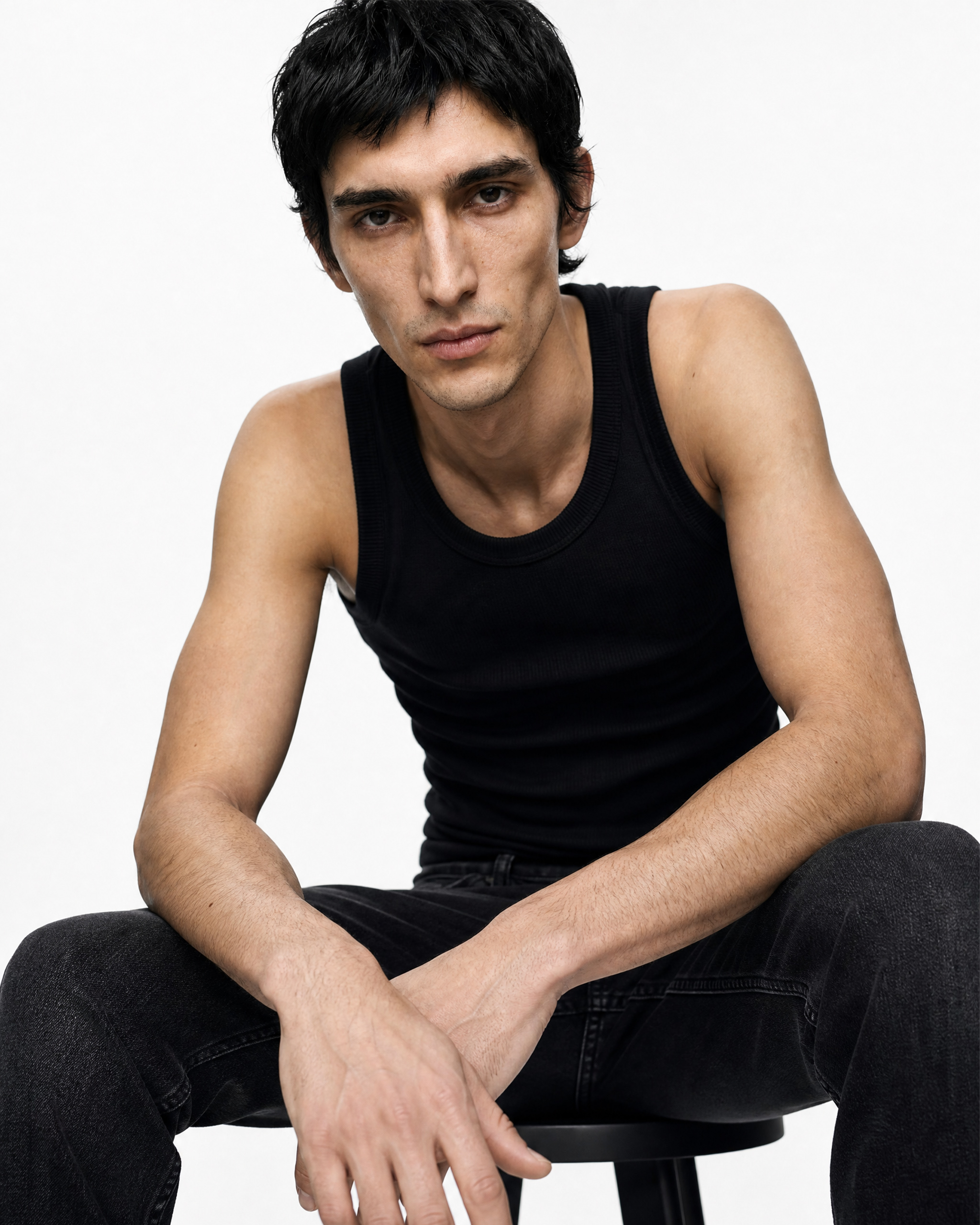 Man in a black sleeveless shirt sits on a stool, facing the camera with a neutral expression.